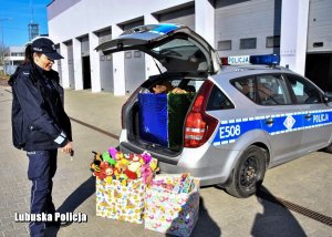 Policjantka przed radiowozem pokazuje na zebrane dla dzieci zabawki