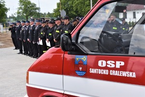 Wóz strażacki, a w tle stojący w szeregu policjanci i strażacy.