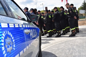 Policyjny radiowóz, a w tle strażacy stojący w szeregu.