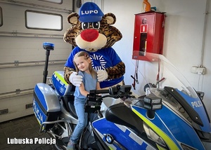 Dziewczynka siedząca na policyjnym motocyklu w obecności maskotki Lubuskiej Policji.
