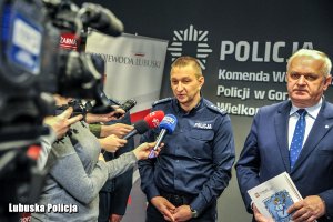 Policjant przemawia do mikrofonu, obok inny mężczyzna.