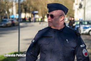 Policjant w mundurze, a w tle policyjny radiowóz.