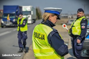 Policjantka drogówki przy kontrolowanym pojeździe, a w tle jadące pojazdy i stojący policjanci.