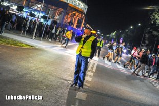 Policjant drogówki na skrzyżowaniu kieruje ruchem, w tle kibice opuszczający stadion.