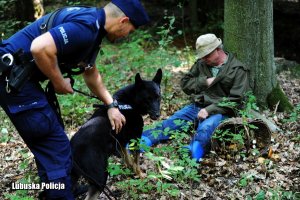 Policjant z psem służbowym przy mężczyźnie siedzącym przy drzewie w lesie.