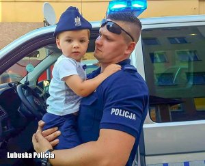 Policjant trzyma dziecko na rękach przy radiowozie.