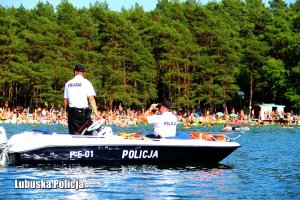 Policjanci na motorówce patrzą w kierunku brzegu jeziora. W tle osoby na plaży.