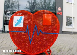 Czerwony, metalowy pojemnik w kształcie serca, a w nim plastikowe nakrętki - w tle budynek komendy Policji.