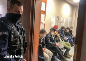 Policjant na korytarzu w komendzie, a w pomieszczeniu mężczyźni siedzący na krzesłach - twarze mężczyzn zamazane graficznie.