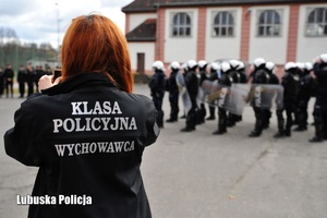 Kobieta z kurtka z napisem klasa policyjna. W tle policjanci oddziałów prewencji