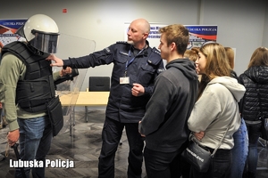 Grupa licealistów rozmawia z policjantem i przymierza wyposażenie policjanta oddziałów prewencji