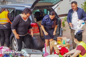 Policjantka z innymi osobami pakuje przedmioty zebrane podczas zbiórki do samochodu.