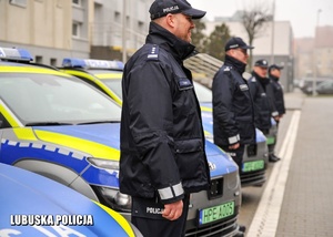 Policjanci stojący w rzędzie obok zaparkowanych radiowozów.