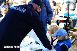 Policjant, a obok niego kobieta z dzieckiem na policyjnym stoisku promocyjnym.