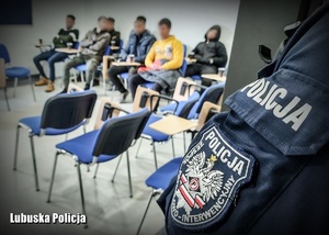 Nielegalni imigranci siedzący na sali odpraw, a na pierwszym planie policjant.