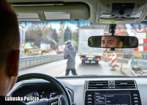 Policjant we wnętrzu radiowozu przed którym osoba piesza przechodzie przez jezdnię.