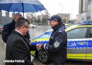 Policjant odbiera kluczyki od mężczyzny podczas uroczystości przekazania nowego radiowozu.