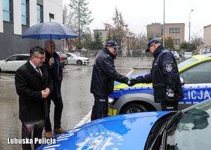 Policjant odbiera gratulacje od zastępcy komendanta wojewódzkiego Policji podczas uroczystości przekazania nowego radiowozu.