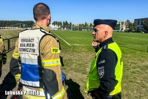 Zastępca Komenda Powiatowego Policji w Nowej Soli rozmawia ze strażakiem na boisku piłkarskim.