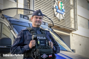 Policjant pozujący do zdjęcia na tle radiowozu i budynku Policji.