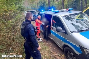 Starszy mężczyzna z ratownikiem medycznym oraz policjantami przy radiowozie.