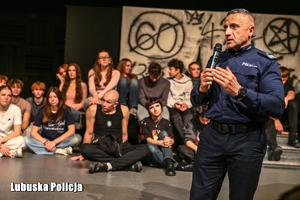 policjant przemawia a za nim na scenie siedzą osoby