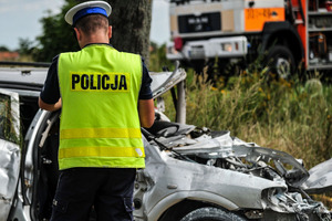 Miejsce wypadku drogowego - rozbity pojazd i pracujący na miejscu policjant.